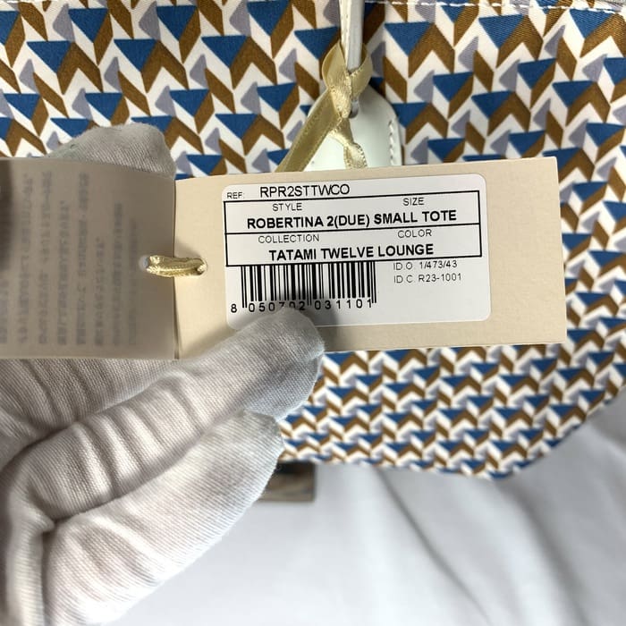 تحمل اليد التي ترتدي القفاز علامة المنتج "ROBERTINA 2 (DUE) SMALL TOTE" فوق قماش بنمط حصير مع نمط هندسي باللونين الأزرق والبني والأبيض ، وهو النمط المميز لمجموعة حمل روبرتا بييري الصغيرة.