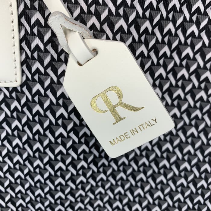 بطاقة ROBERTA PIERI الجلدية البيضاء مع الأحرف "R" باللون الذهبي وكلمات "MADE IN ITALY" تتدلى من خلفية قماش هندسي بالأبيض والأسود.