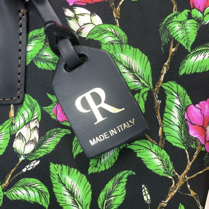 بطاقة الأمتعة ROBERTA PIERI السوداء مع الحرف "R" الذهبي والنص الإيطالي معلقة من قماش زهري بأوراق خضراء وزهور وردية ، تكمل القماش الخشن الصغير بأناقة إيطالية خالدة.