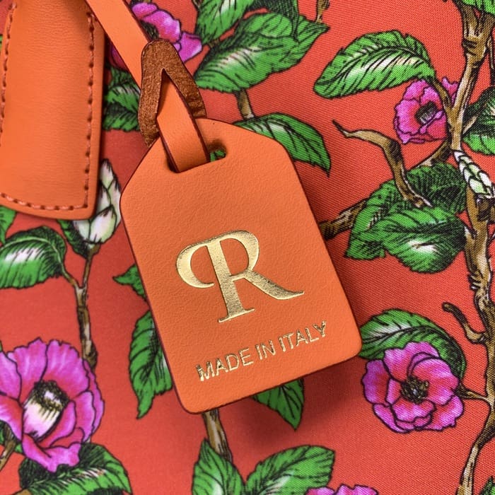 لقطة مقربة للبطاقة الجلدية البرتقالية "ROBERTA PIERI" مع حرف R ذهبي ونص إيطالي، مثبتة على قماش زهري أحمر مزين بزهور وردية وأوراق خضراء على حقيبة دفل صغيرة.